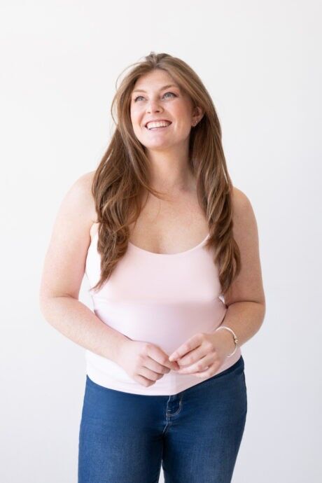 A woman wearing a pink tank top and jeans, showcasing a scoop neck spaghetti strap layering camisole